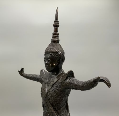 fig 4. A dark bronze sculpture depicting a classical court dancer in ceremonial attire with a tall spired headdress, balanced on one foot atop an octagonal stepped base.