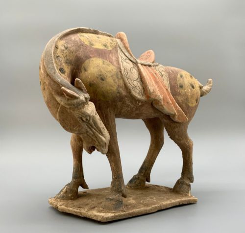 fig 3. A terracotta horse figurine featuring a dappled coat pattern, draped saddle, and a head lowered towards its front hoof on a rectangular plinth.