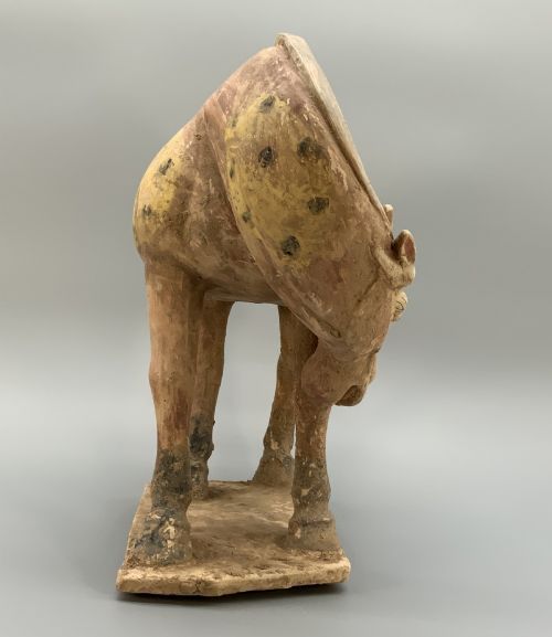 fig 5. A terracotta horse figurine featuring a dappled coat pattern, draped saddle, and a head lowered towards its front hoof on a rectangular plinth.