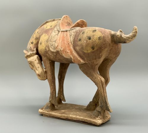 fig 6. A terracotta horse figurine featuring a dappled coat pattern, draped saddle, and a head lowered towards its front hoof on a rectangular plinth.