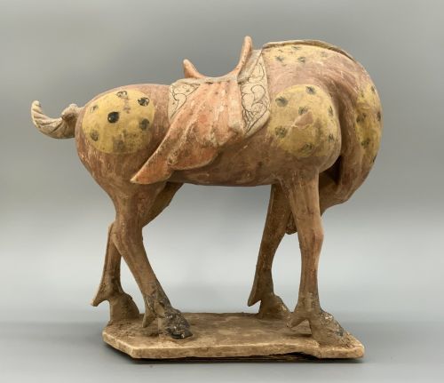 fig 7. A terracotta horse figurine featuring a dappled coat pattern, draped saddle, and a head lowered towards its front hoof on a rectangular plinth.