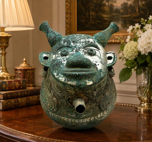 fig 1. A cast bronze ritual vessel featuring a stylised human face with horn-like projections, a small tubular spout, and textured surface oxidisation.