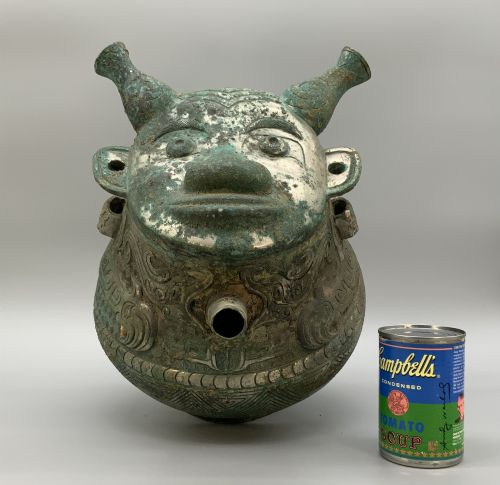 fig 2. A cast bronze ritual vessel featuring a stylised human face with horn-like projections, a small tubular spout, and textured surface oxidisation.