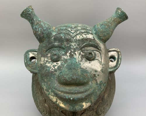fig 3. A cast bronze ritual vessel featuring a stylised human face with horn-like projections, a small tubular spout, and textured surface oxidisation.