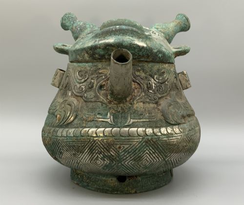fig 4. A cast bronze ritual vessel featuring a stylised human face with horn-like projections, a small tubular spout, and textured surface oxidisation.