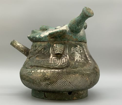 fig 6. A cast bronze ritual vessel featuring a stylised human face with horn-like projections, a small tubular spout, and textured surface oxidisation.