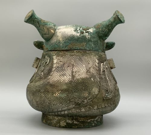 fig 7. A cast bronze ritual vessel featuring a stylised human face with horn-like projections, a small tubular spout, and textured surface oxidisation.