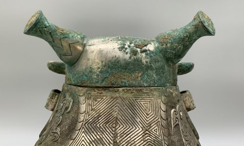 fig 8. A cast bronze ritual vessel featuring a stylised human face with horn-like projections, a small tubular spout, and textured surface oxidisation.