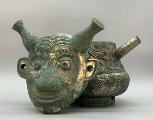 fig 9. A cast bronze ritual vessel featuring a stylised human face with horn-like projections, a small tubular spout, and textured surface oxidisation.