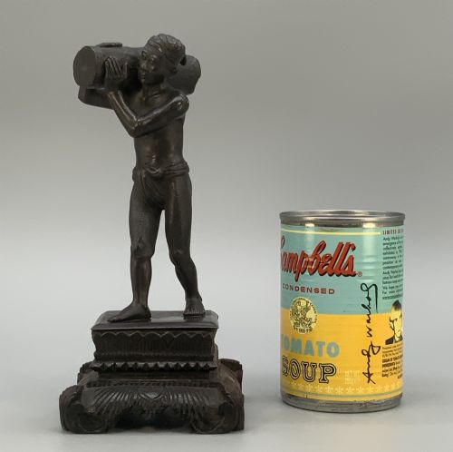 fig 2. A dark patinated bronze figurine of a man carrying a log on his shoulder, mounted on an intricately carved wooden stand.