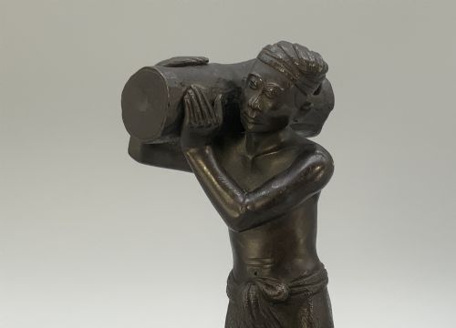 fig 3. A dark patinated bronze figurine of a man carrying a log on his shoulder, mounted on an intricately carved wooden stand.