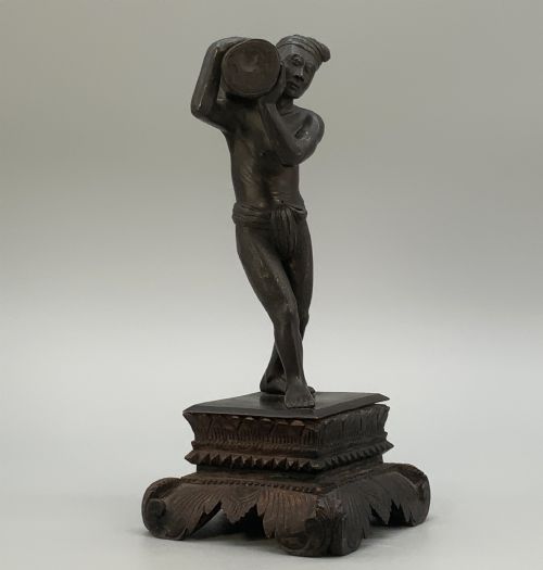 fig 4. A dark patinated bronze figurine of a man carrying a log on his shoulder, mounted on an intricately carved wooden stand.