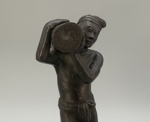 fig 5. A dark patinated bronze figurine of a man carrying a log on his shoulder, mounted on an intricately carved wooden stand.