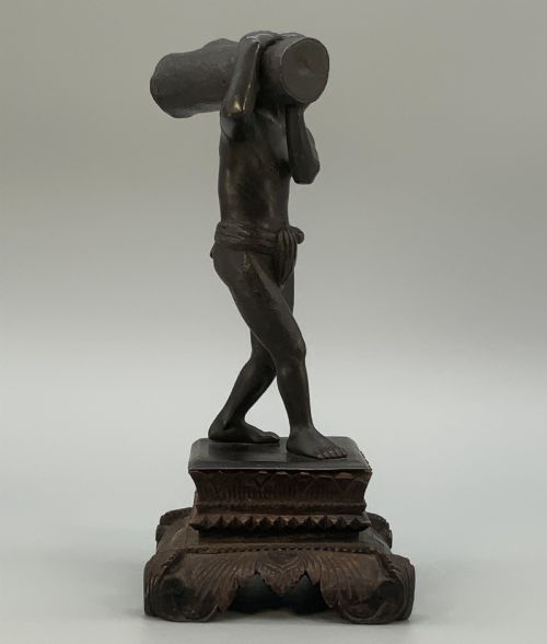 fig 7. A dark patinated bronze figurine of a man carrying a log on his shoulder, mounted on an intricately carved wooden stand.