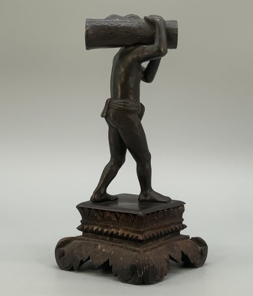 fig 8. A dark patinated bronze figurine of a man carrying a log on his shoulder, mounted on an intricately carved wooden stand.