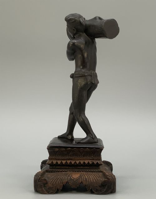 fig 10. A dark patinated bronze figurine of a man carrying a log on his shoulder, mounted on an intricately carved wooden stand.