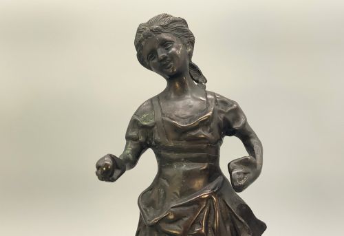 fig 3. An antique dark bronze sculpture of a young maiden wearing a draped apron dress, standing barefoot on a rounded integral base.