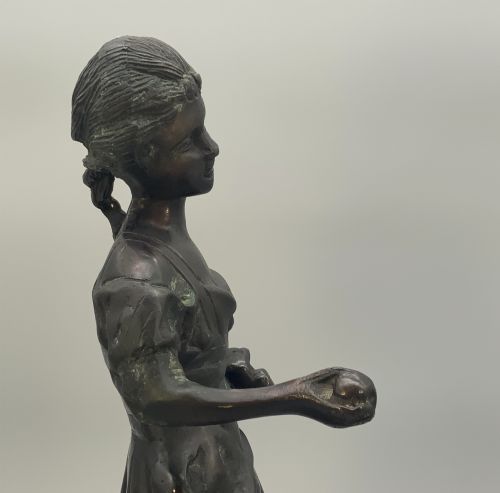 fig 9. An antique dark bronze sculpture of a young maiden wearing a draped apron dress, standing barefoot on a rounded integral base.