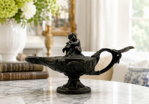 fig 1. A patinated dark bronze classical oil lamp featuring a seated putto figure on top and a scroll handle resting on a pedestal base.
