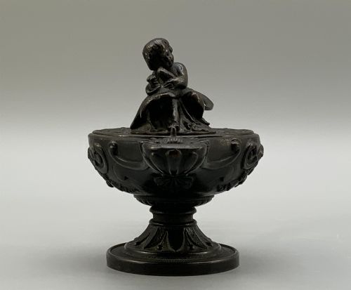 fig 3. A patinated dark bronze classical oil lamp featuring a seated putto figure on top and a scroll handle resting on a pedestal base.