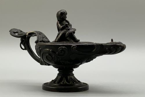 fig 4. A patinated dark bronze classical oil lamp featuring a seated putto figure on top and a scroll handle resting on a pedestal base.