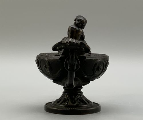 fig 5. A patinated dark bronze classical oil lamp featuring a seated putto figure on top and a scroll handle resting on a pedestal base.