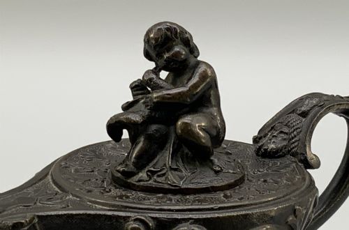 fig 6. A patinated dark bronze classical oil lamp featuring a seated putto figure on top and a scroll handle resting on a pedestal base.