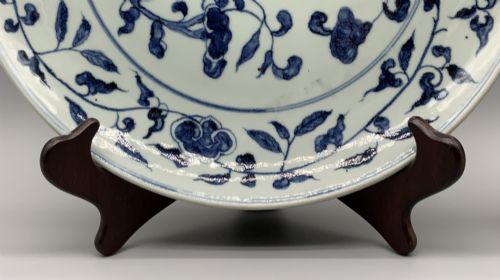 fig 6. A circular Chinese blue and white porcelain charger decorated with hand-painted scrolling foliage and floral motifs, resting on a dark wooden stand.
