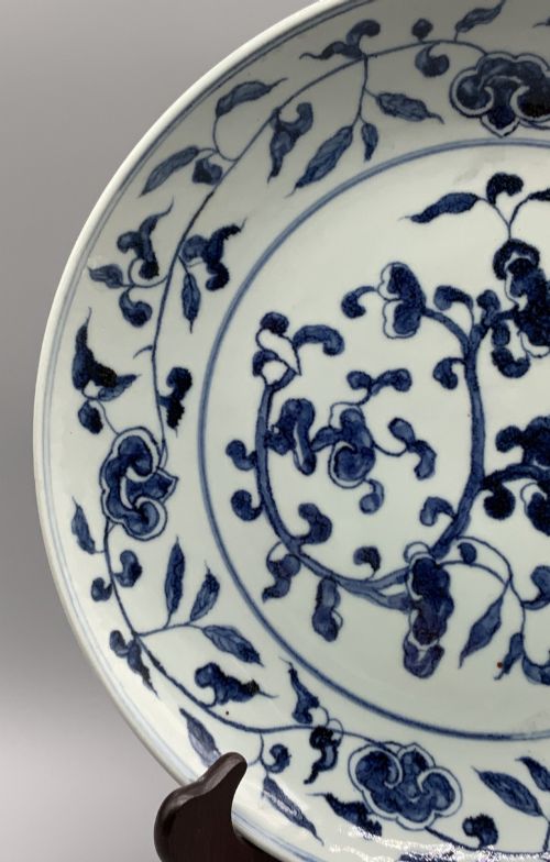 fig 8. A circular Chinese blue and white porcelain charger decorated with hand-painted scrolling foliage and floral motifs, resting on a dark wooden stand.