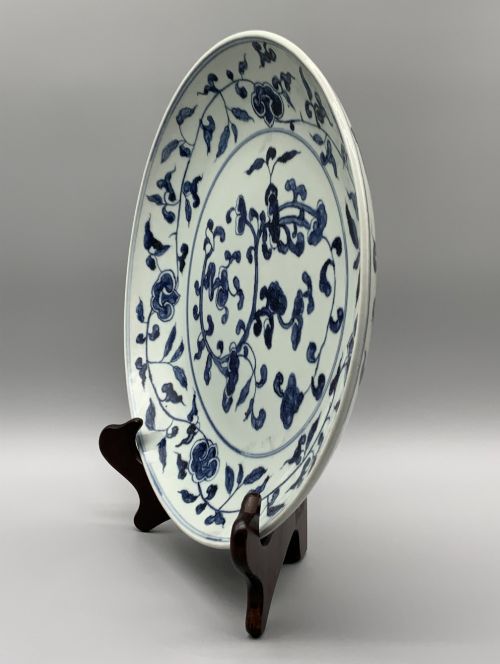 fig 9. A circular Chinese blue and white porcelain charger decorated with hand-painted scrolling foliage and floral motifs, resting on a dark wooden stand.