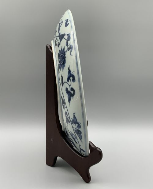fig 10. A circular Chinese blue and white porcelain charger decorated with hand-painted scrolling foliage and floral motifs, resting on a dark wooden stand.