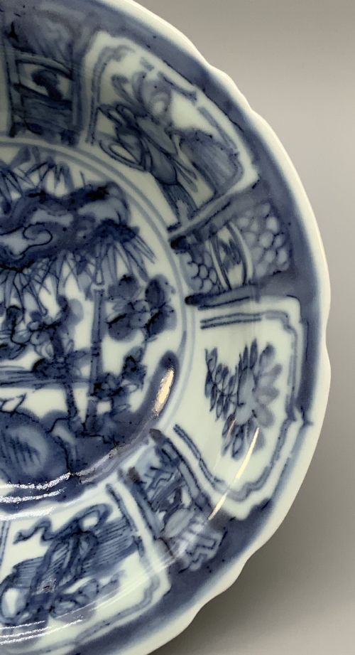fig 6. A circular porcelain bowl with a lobed edge, decorated in cobalt blue underglaze featuring a central landscape surrounded by panelled floral reserves.