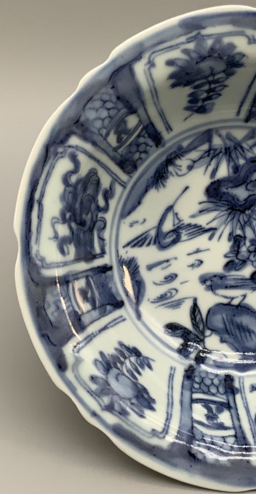 fig 7. A circular porcelain bowl with a lobed edge, decorated in cobalt blue underglaze featuring a central landscape surrounded by panelled floral reserves.