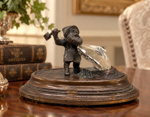 fig 1. A small dark bronze figurine of a bearded dwarf miner holding a hammer overhead, standing on a molded base next to a faceted cut crystal gem.