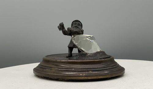 fig 3. A small dark bronze figurine of a bearded dwarf miner holding a hammer overhead, standing on a molded base next to a faceted cut crystal gem.