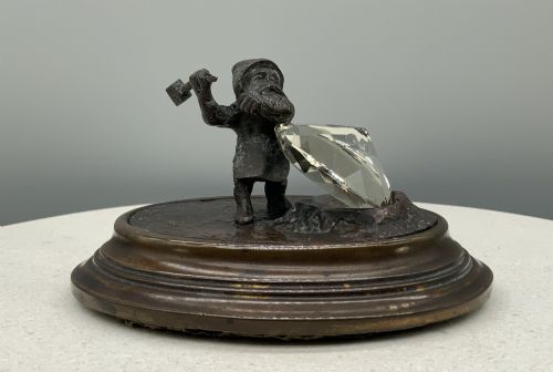 fig 4. A small dark bronze figurine of a bearded dwarf miner holding a hammer overhead, standing on a molded base next to a faceted cut crystal gem.