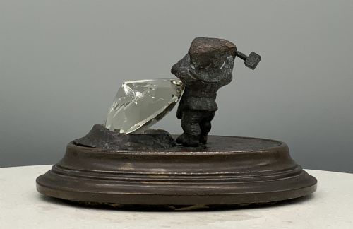 fig 6. A small dark bronze figurine of a bearded dwarf miner holding a hammer overhead, standing on a molded base next to a faceted cut crystal gem.