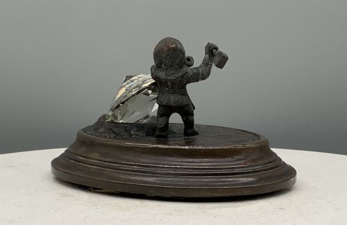 fig 7. A small dark bronze figurine of a bearded dwarf miner holding a hammer overhead, standing on a molded base next to a faceted cut crystal gem.