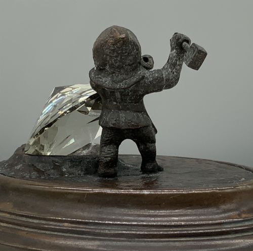 fig 8. A small dark bronze figurine of a bearded dwarf miner holding a hammer overhead, standing on a molded base next to a faceted cut crystal gem.