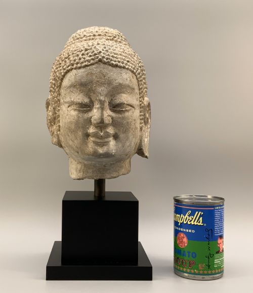 fig 2. A carved limestone Chinese Buddha head featuring downcast eyes, elongated earlobes, and coiled hair, mounted on a black square stand.