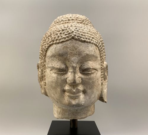fig 3. A carved limestone Chinese Buddha head featuring downcast eyes, elongated earlobes, and coiled hair, mounted on a black square stand.