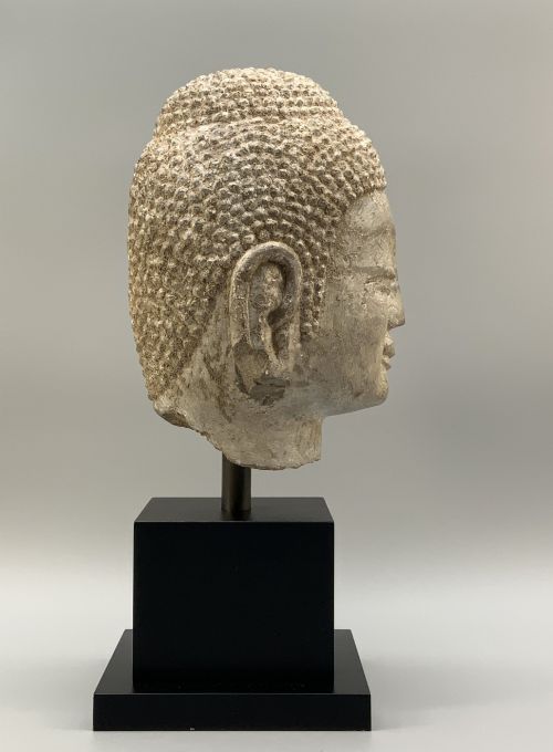 fig 4. A carved limestone Chinese Buddha head featuring downcast eyes, elongated earlobes, and coiled hair, mounted on a black square stand.
