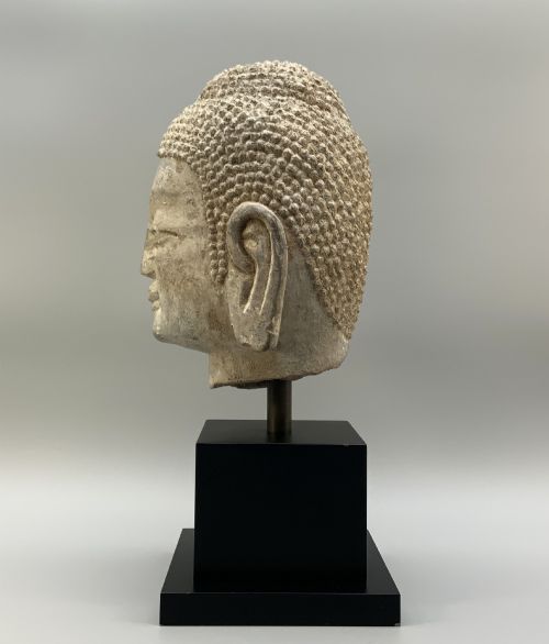 fig 6. A carved limestone Chinese Buddha head featuring downcast eyes, elongated earlobes, and coiled hair, mounted on a black square stand.