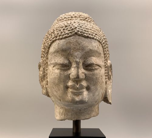 fig 9. A carved limestone Chinese Buddha head featuring downcast eyes, elongated earlobes, and coiled hair, mounted on a black square stand.