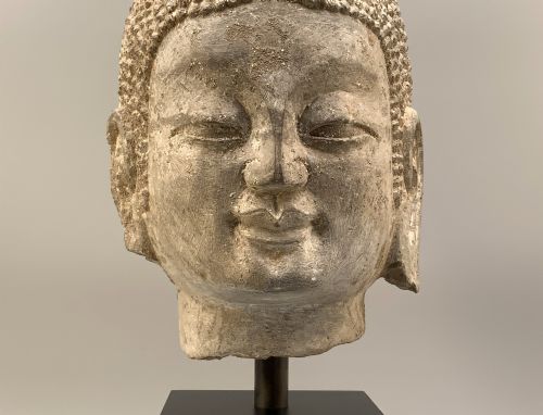 fig 10. A carved limestone Chinese Buddha head featuring downcast eyes, elongated earlobes, and coiled hair, mounted on a black square stand.