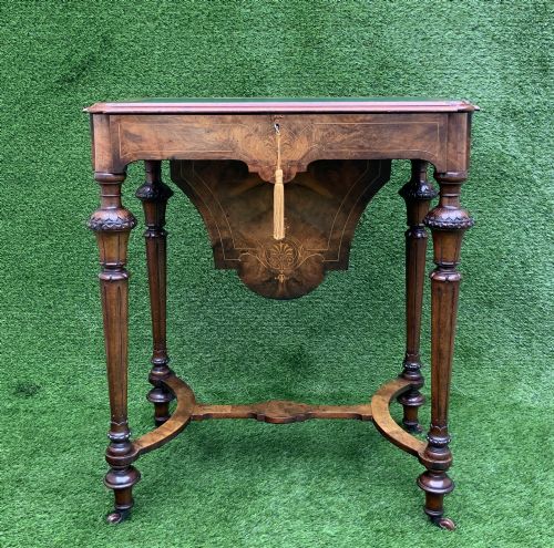 fig 1. A 19th-century burr walnut work table featuring a green leather inset top, floral marquetry inlay, turned legs, and a hanging storage pouch.