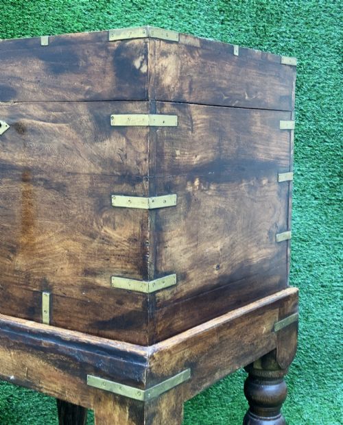 fig 7. A dark teak Anglo-Indian wine cellarette box with brass corner mounts resting on a carved turned-leg wooden stand.