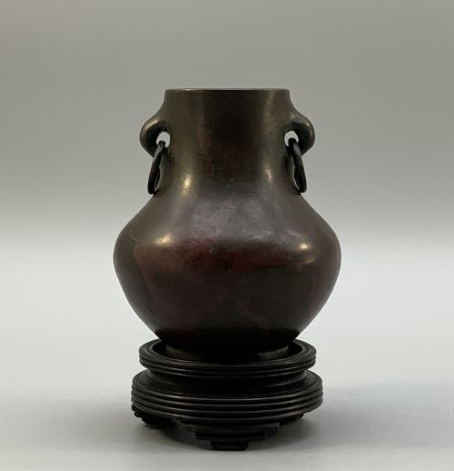 fig 1. A small pear-shaped dark bronze vase with loop handles and loose rings, featuring mottled red surface patches, resting on a carved dark wooden base.
