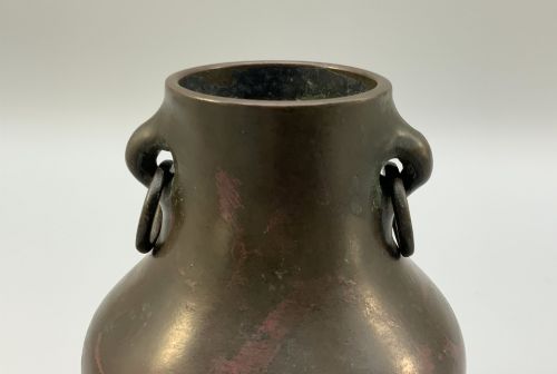 fig 3. A small pear-shaped dark bronze vase with loop handles and loose rings, featuring mottled red surface patches, resting on a carved dark wooden base.