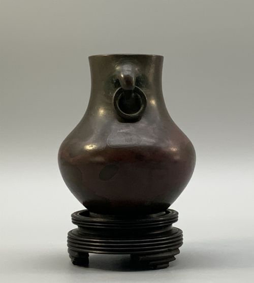 fig 5. A small pear-shaped dark bronze vase with loop handles and loose rings, featuring mottled red surface patches, resting on a carved dark wooden base.